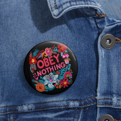 A black round pinback button featuring floral embroidery-style designs and bold red text that reads "Obey Nothing."